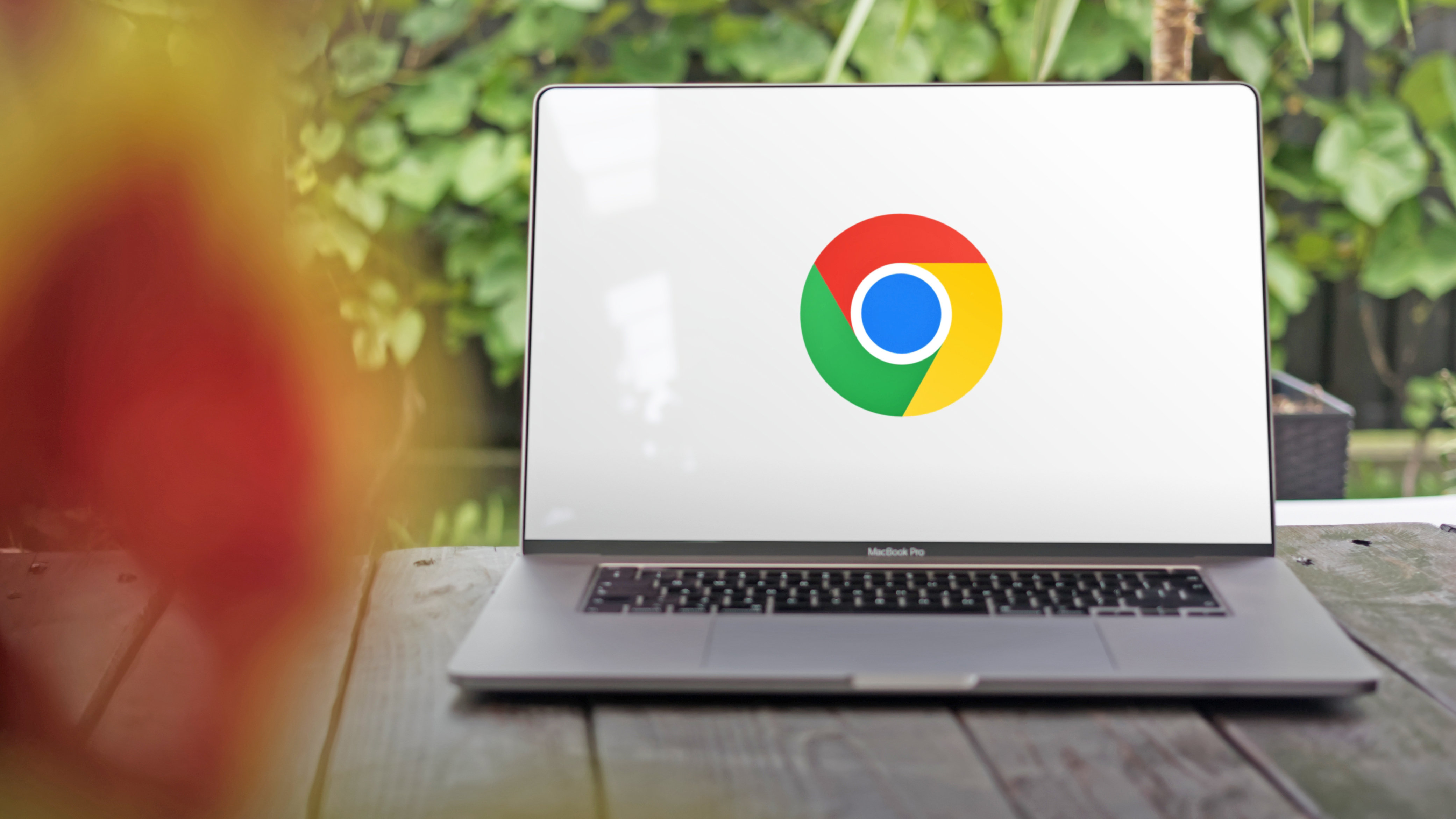 Arkasında yeşil yapraklar bulunan bir masanın üzerinde duran büyük bir MacBook Pro'nun ekranında, beyaz bir arka plan üzerinde Google Chrome logosunun yer aldığı bir fotoğraf.