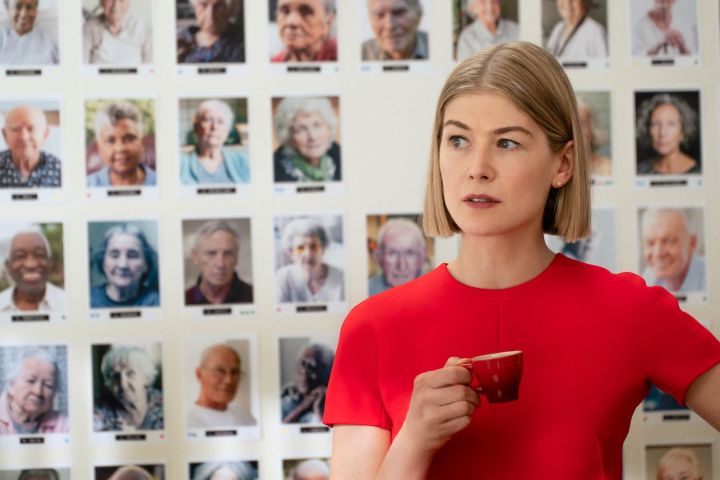 Rosamund Pike, fotoğraflardan oluşan bir duvarın önünde bir fincan kahve tutuyor.