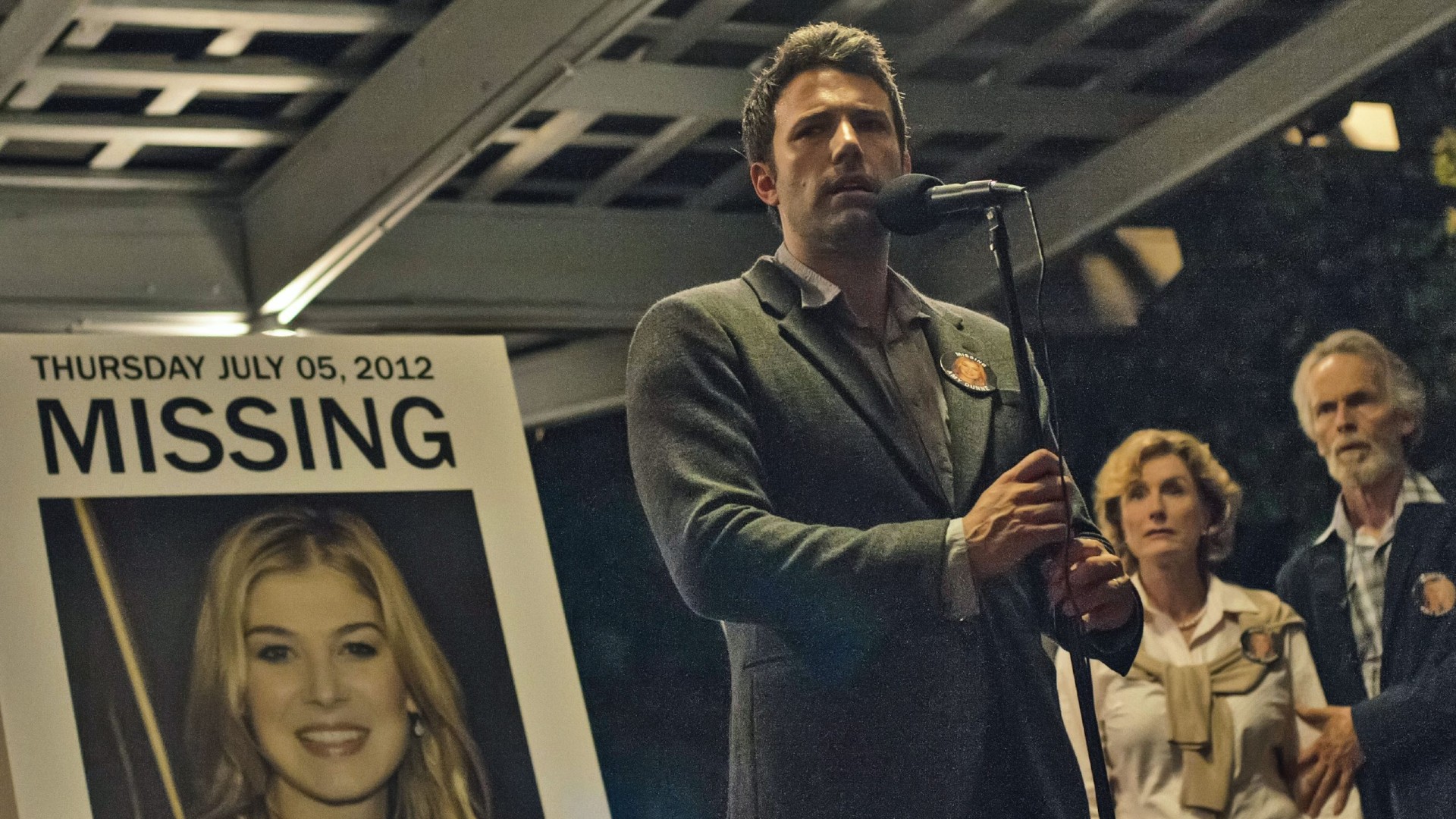 Ben Affleck, Gone Girl filminden bir karede Rosamund Pike'ın elinde mikrofonla büyütülmüş bir fotoğrafının önünde duruyor.