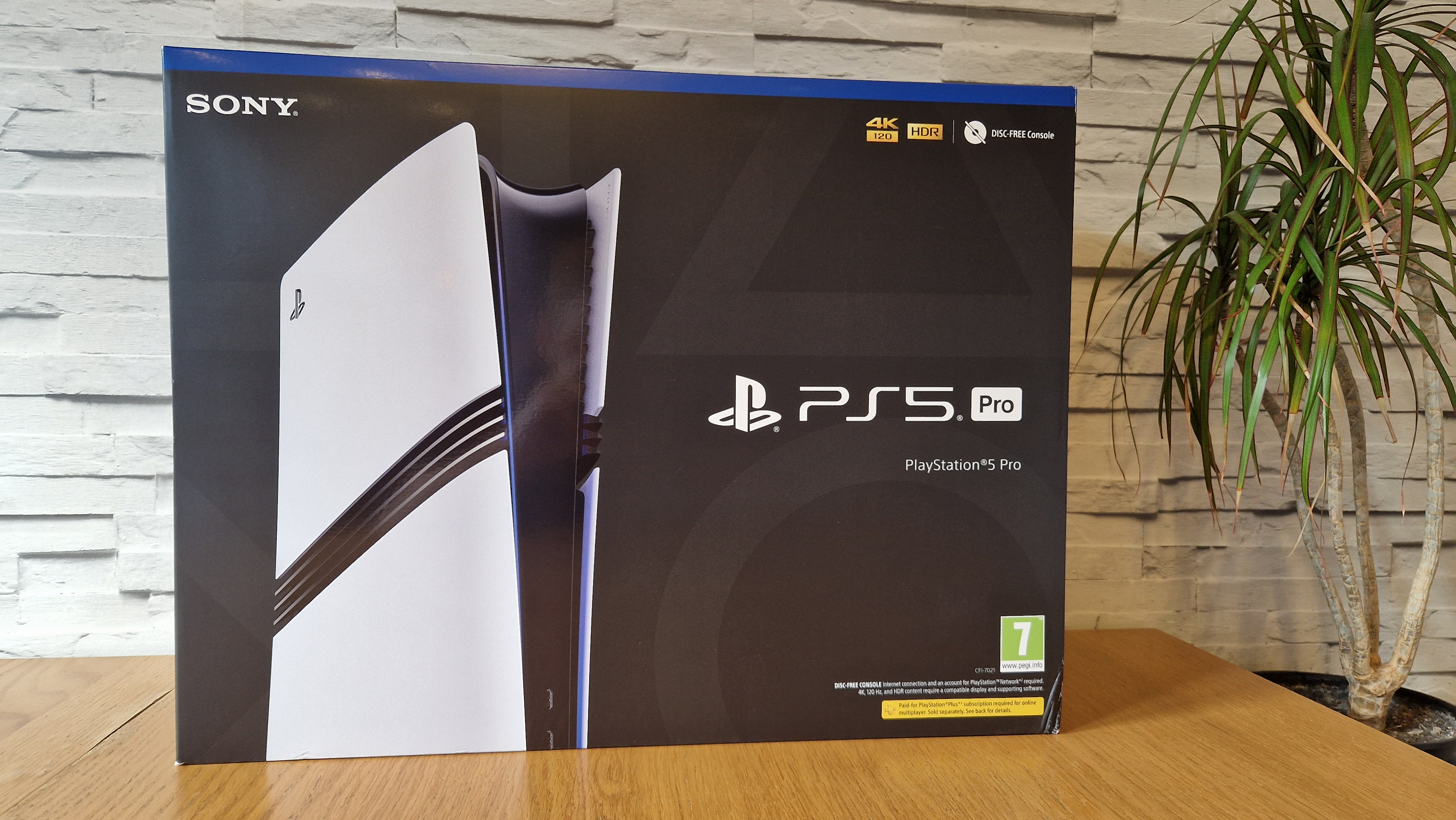 Beyaz tuğlalı bir arka planın önünde ve saksı bitkisinin yanında ahşap bir masanın üzerinde bulunan PS5 Pro kutusu