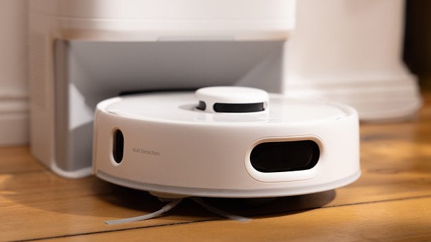Şarj istasyonunun yanındaki ahşap zemin üzerinde, duvar algılama işlevine sahip SwitchBot K10 Pro robot elektrikli süpürge.