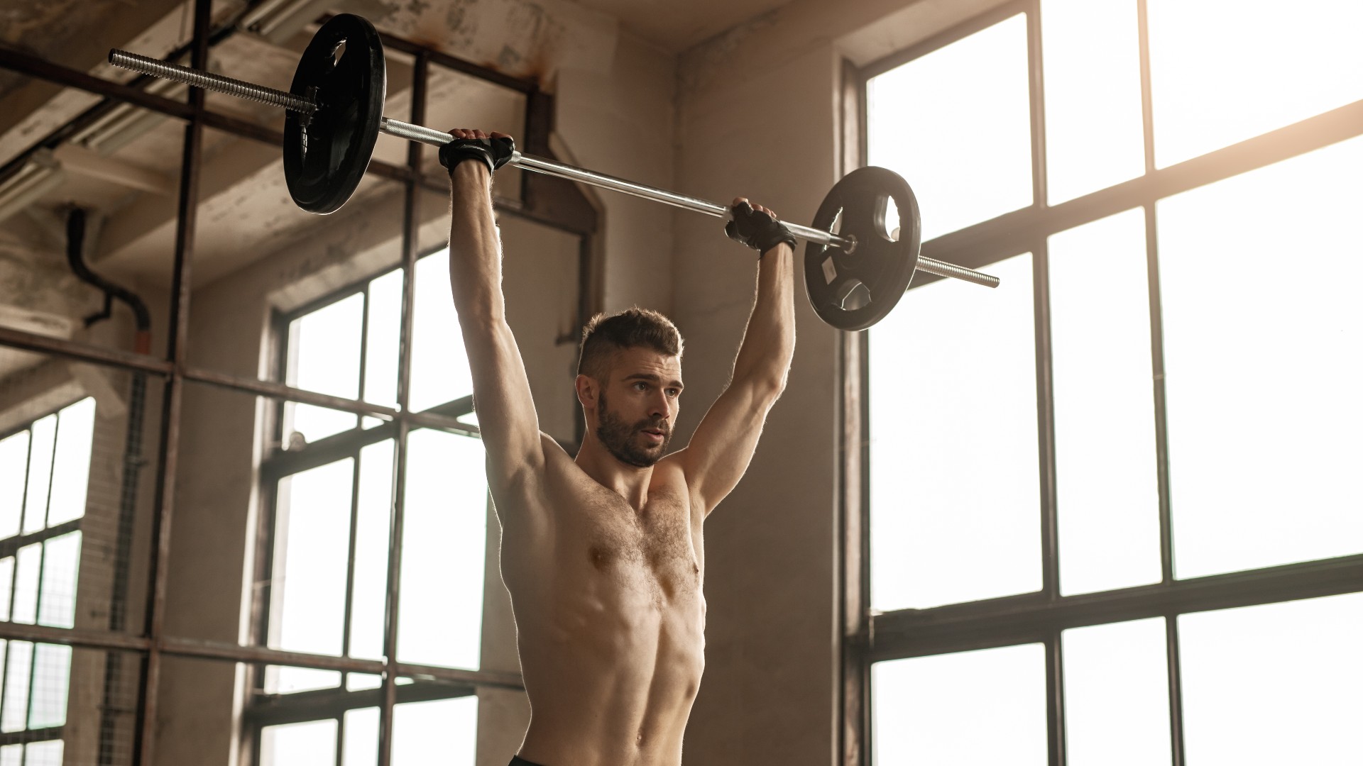 Spor salonunda barı başının üzerine uzatarak halterle baş üstü pres yapan adam