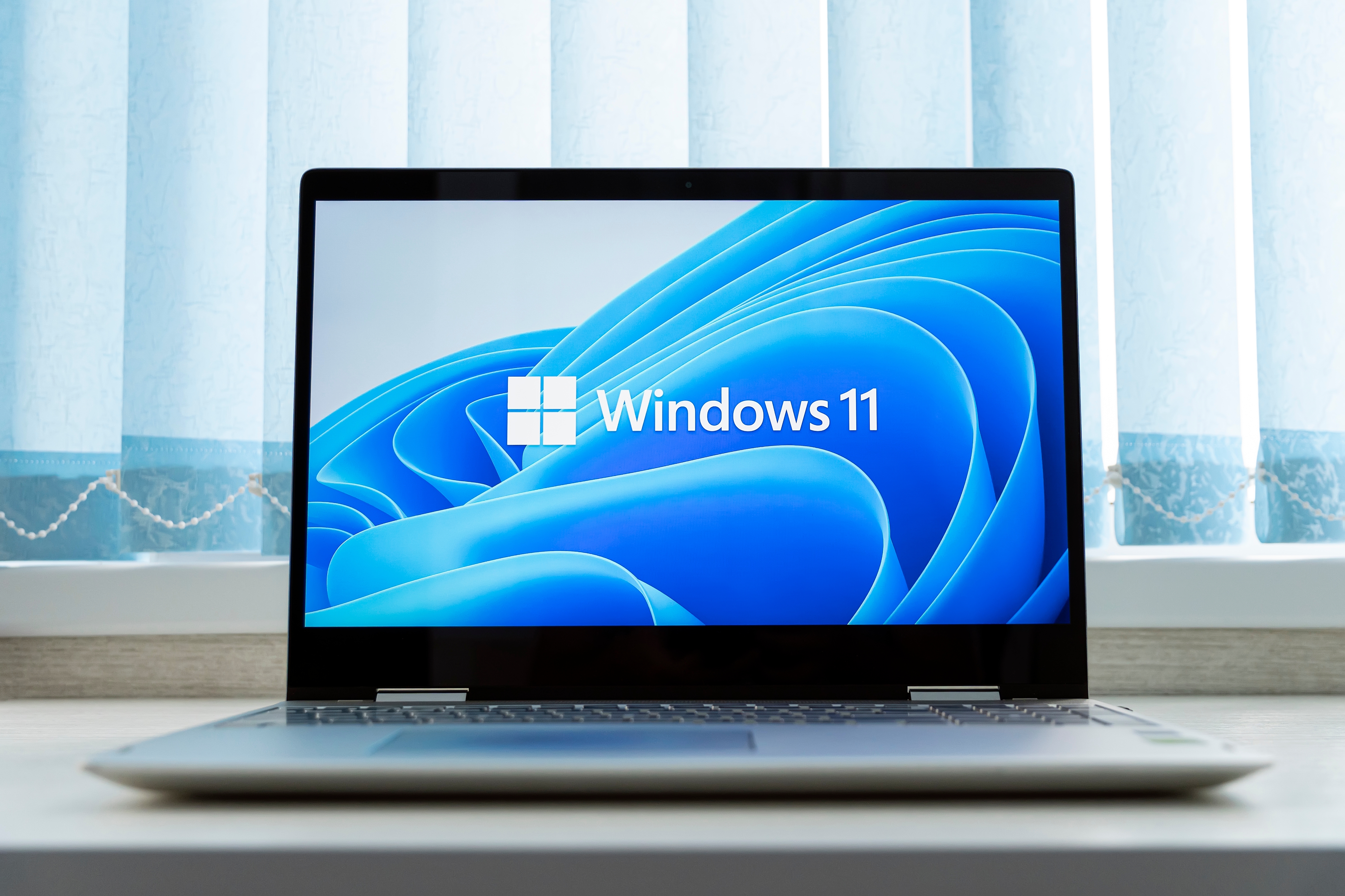 Pencerenin önündeki masanın üzerinde duran Windows 11 dizüstü bilgisayar