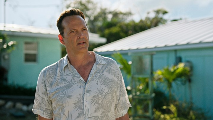 Vince Vaughn çiçekli düğmeli bluzuyla bir plaj evinin yanında duruyor ve Bad Monkey'den bir sahnede sinirlenmiş görünüyor.
