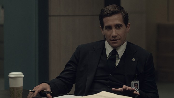 Jake Gyllenhaal, Apple TV+'ta yayınlanan Presumed Innocent programından bir sahnede takım elbise giyerek masanın arkasında oturuyor.