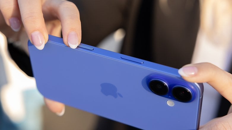 Bir kişi mavi bir iPhone 16 tutuyor ve arkadaki düğmelere ve çift kameraya odaklanıyor.