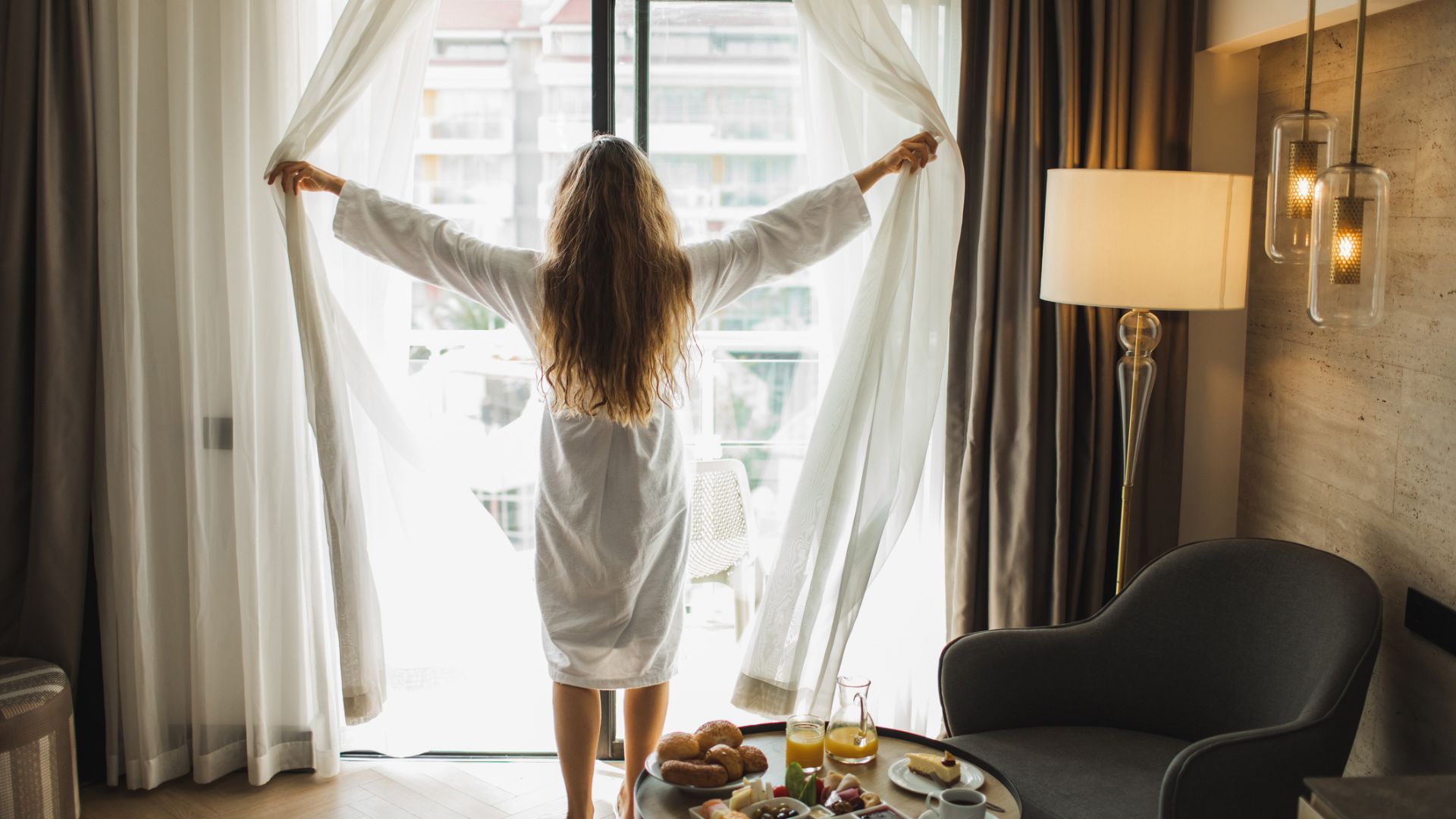 Sabahları bir otel odasının perdelerini açan sabahlıklı bir kadın