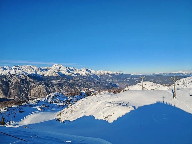 Berrak mavi gökyüzünün altında karla kaplı dağlar ve ön planda kayak teleferikleri var.