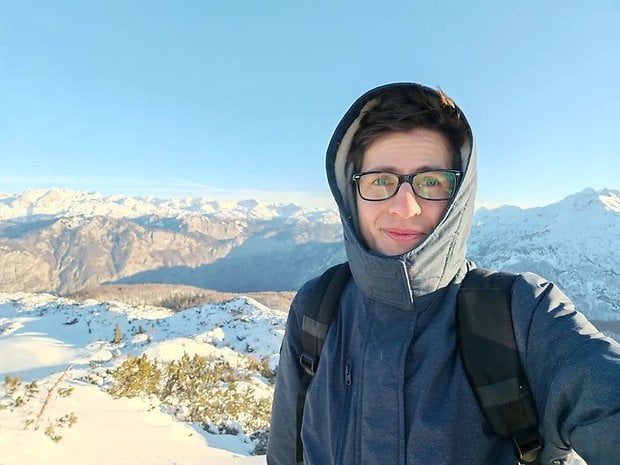 Gözlüklü mavi ceketli bir kişi karla kaplı bir dağ manzarasında açık bir gökyüzünün altında duruyor.