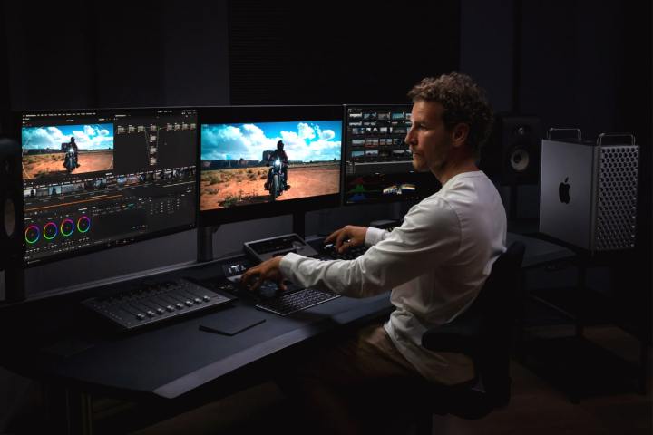 Bir kişi, karanlık bir odada üç monitör ve bir düzenleme konsolunun yanında bir Apple Mac Pro kullanıyor.