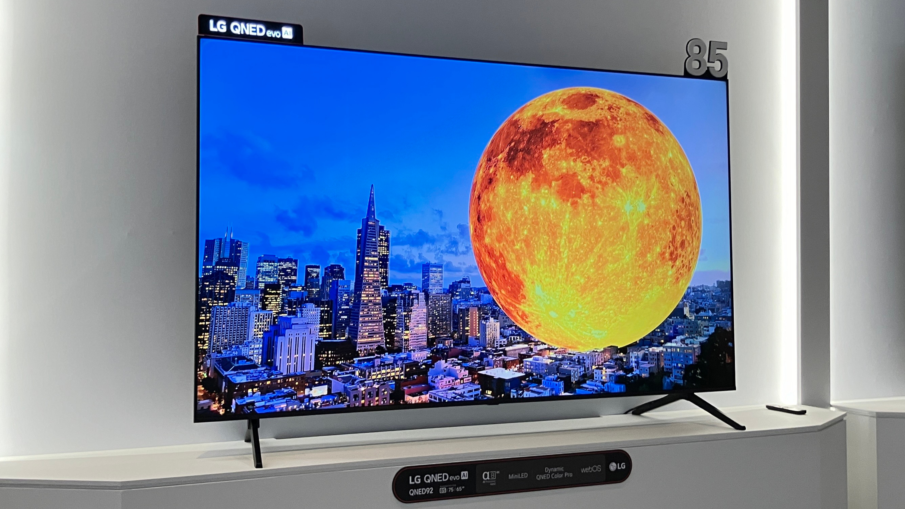 Bir showroom zeminde beyaz renkli bir duvarın önünde sergilenen 85 inç LG QNED92 mini liderliğindeki TV