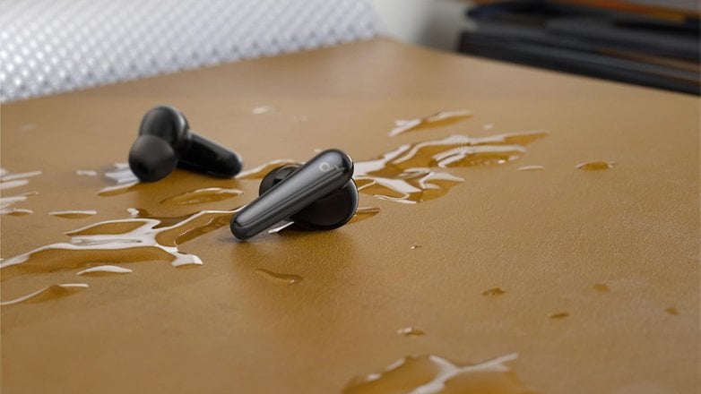 Su damlalarıyla kahverengi bir yüzeyde siyah Anker Soundcore Liberty 4 Pro kulaklık.