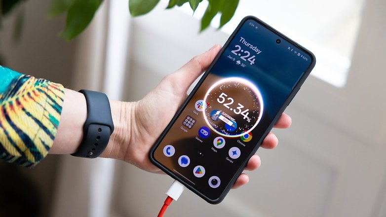 Bir kişi, şarj cihazına bağlı, saati ve pil seviyesini gösteren bir OnePlus 13R akıllı telefon tutuyor.