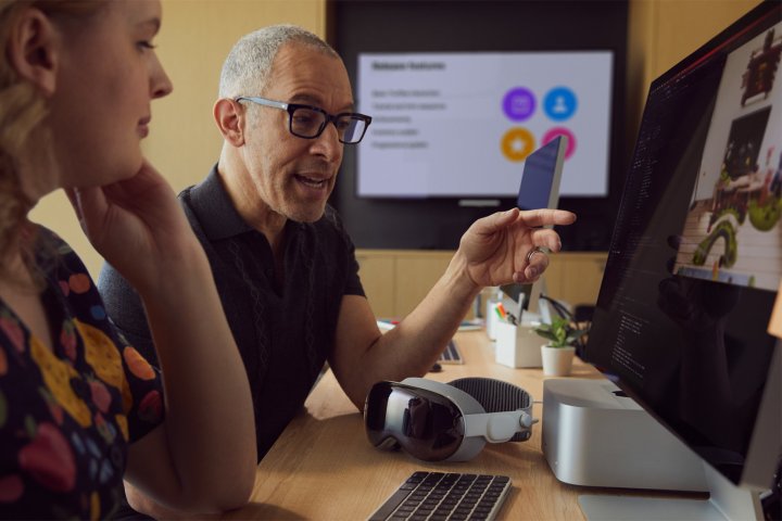 Bir Vision Pro masaya dayanırken bir geliştirici Mac ekranına işaret eder.