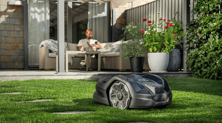 Arka planda bir terasta rahatlayan bir adamla yeşil çimen üzerinde bir Husqvarna 305xe çekirdek çim biçme makinesi. Arka planda bir terasta rahatlayan bir adamla yeşil çimen üzerinde bir Husqvarna 305xe çekirdek çim biçme makinesi.