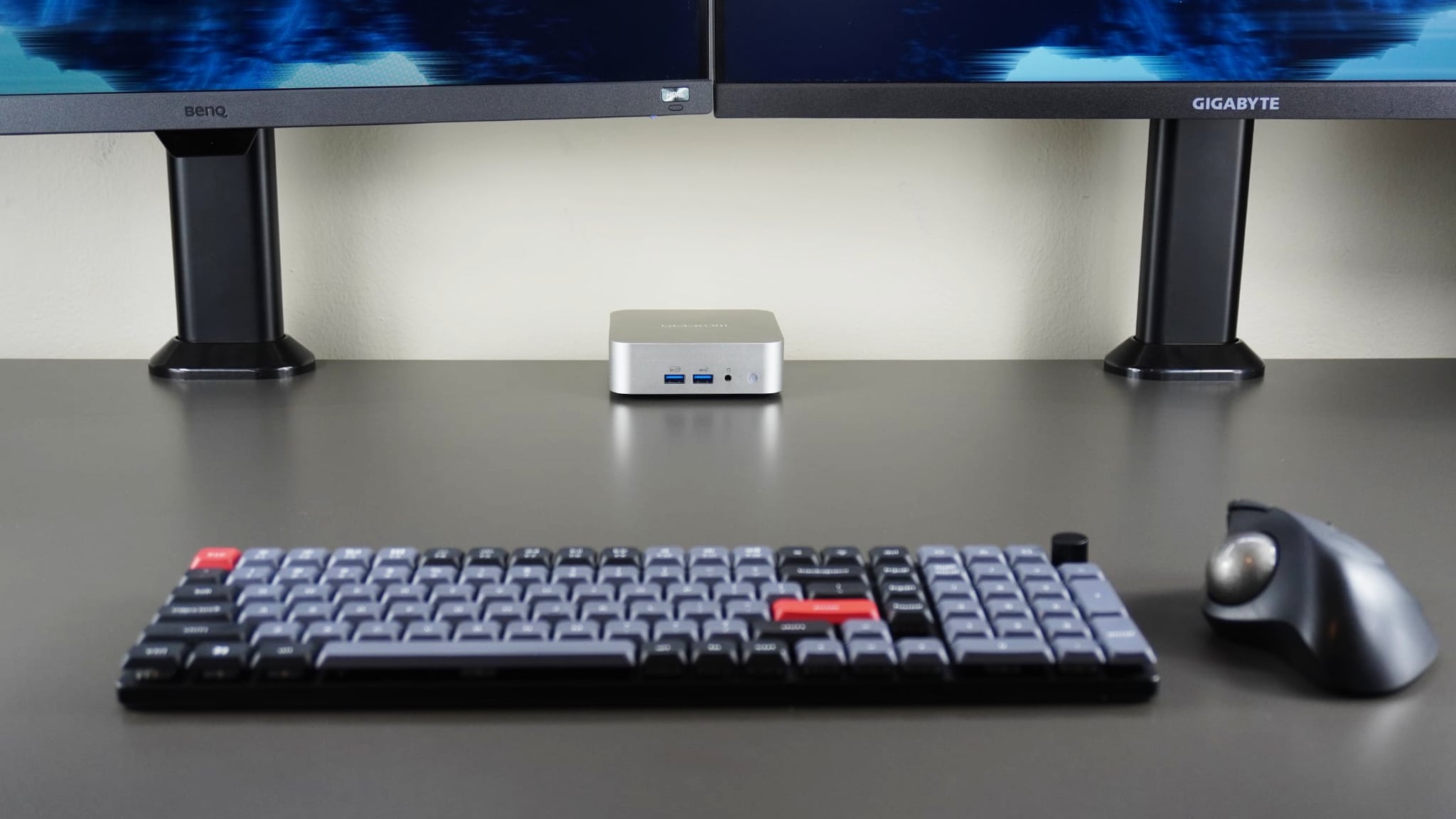 Bir masanın arkasındaki Geekom A6, ön planda mekanik bir klavye ve trackball faresi ile iki monitör altında