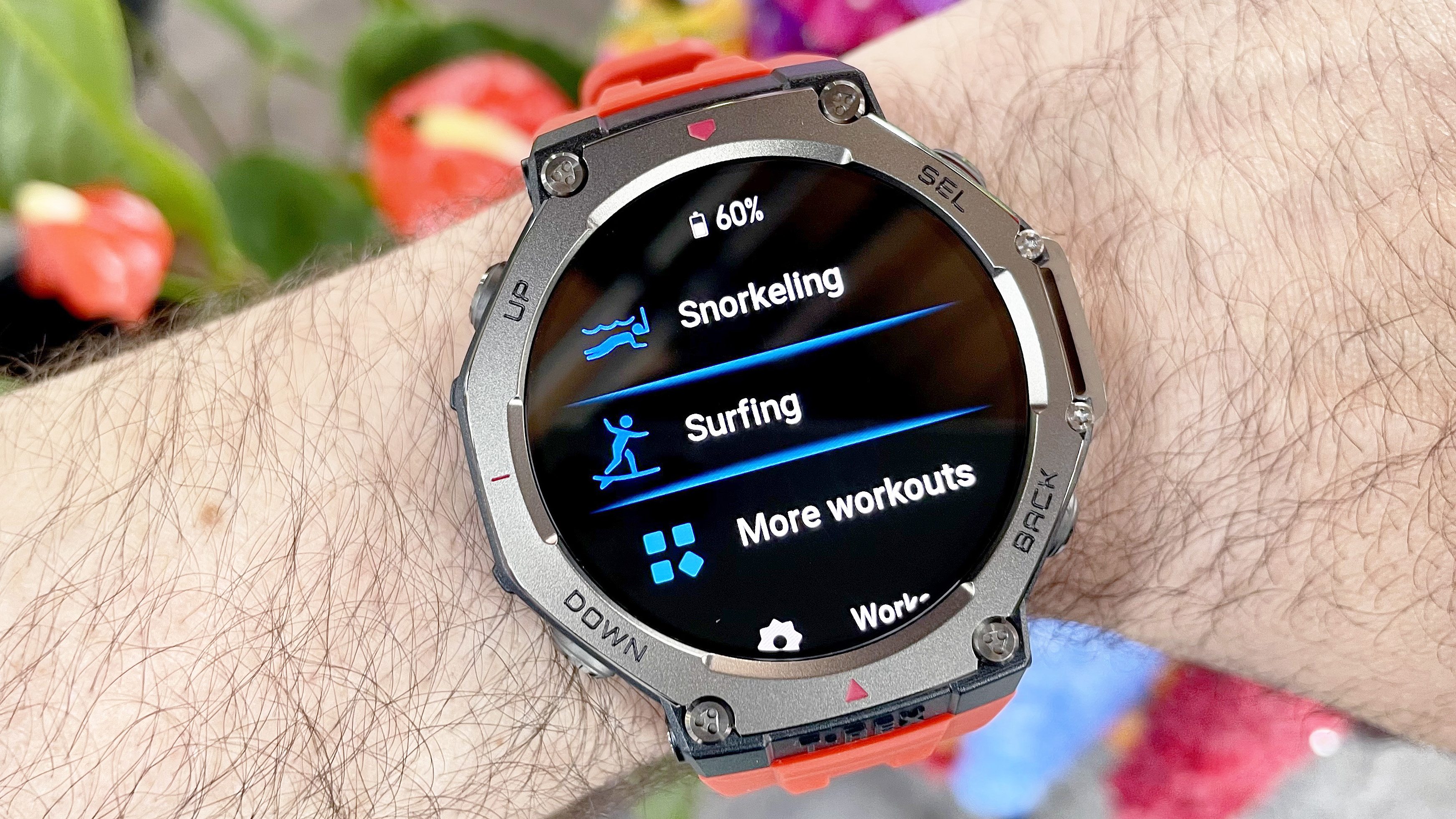 Amazfit T-Rex 3, görüntülenen şnorkel ve sörf egzersiz izleme modları ile bir kullanıcının bileğinde yakın çekim gösterdi; Renkli çiçekler arka planda odak dışı