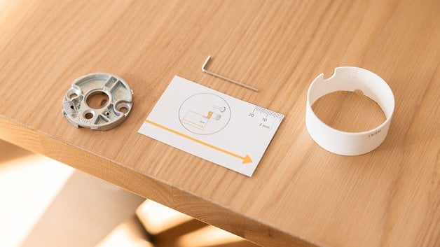 Ahşap bir masada Nuki Smart Lock Pro için bileşenler: metal bir taban, kurulum talimatları ve yuvarlak bir kapak.