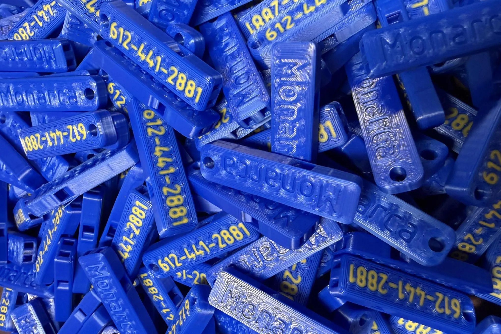 Her bir tarafında “612-441-2881” ve “Monarca” yazılı, mavi 3D yazıcıdan üretilen düdük yığını.