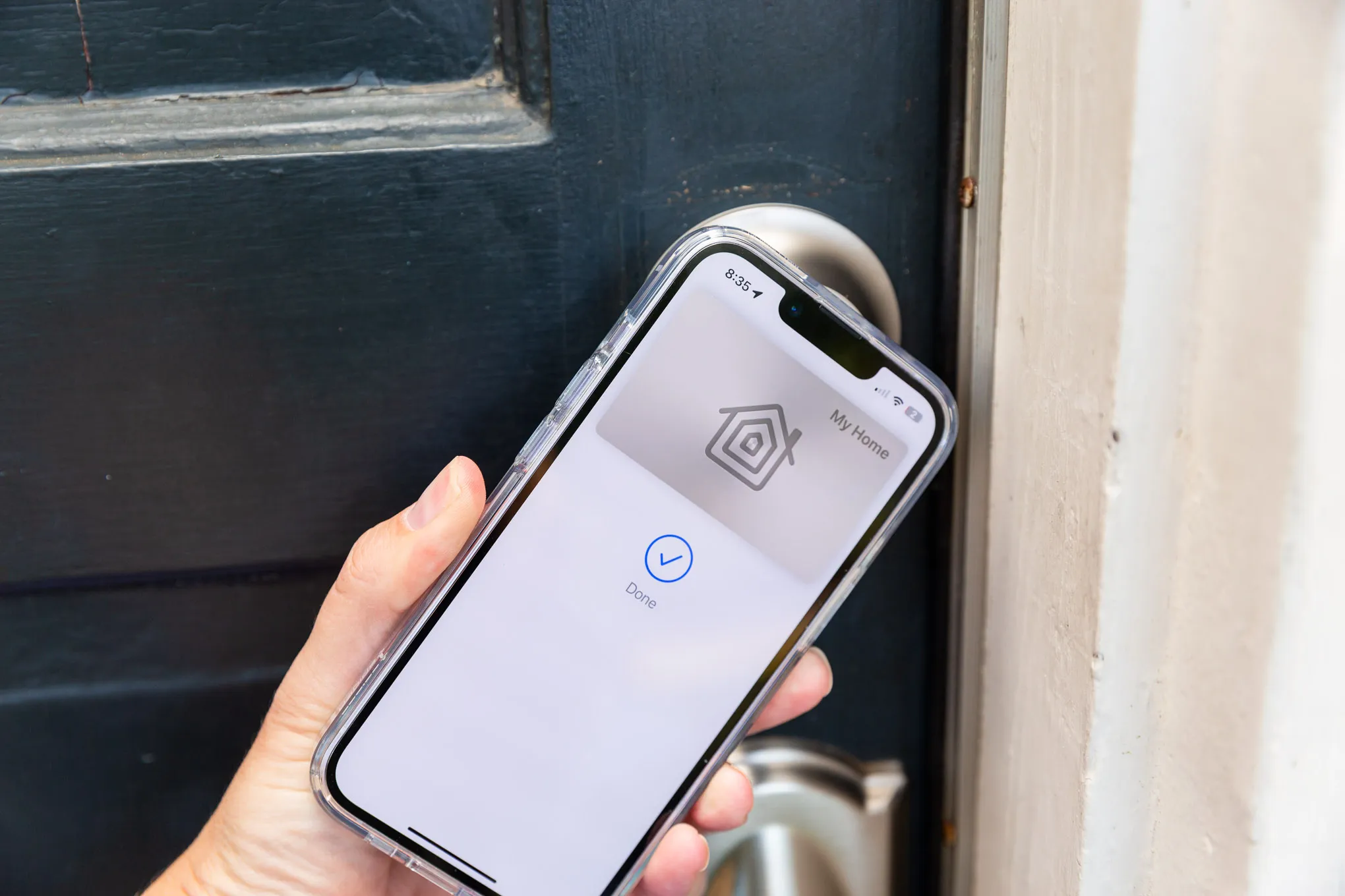 Bir el, bir kapıya iPhone tutuyor. Telefon ekranında bir anahtar kartı görüntüsü var, üstünde Apple Home logosu ve 'Evime' yazısı bu durumda ise mavi bir onay işareti daire içerisinde 'Tamam' yazısı bulunuyor.