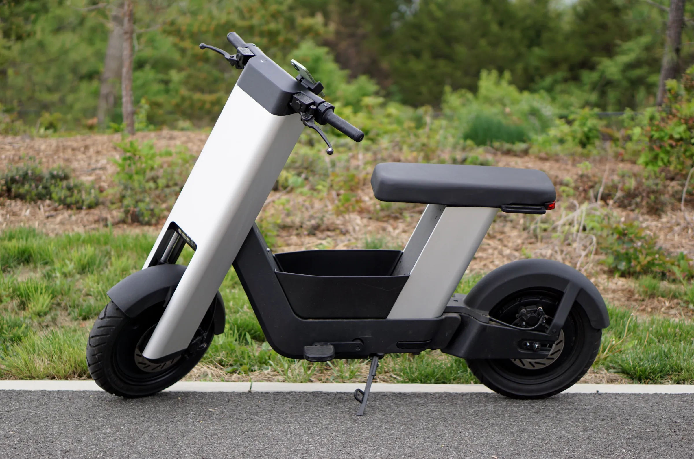A photo of a silver e-bike from the side, in front of trees.